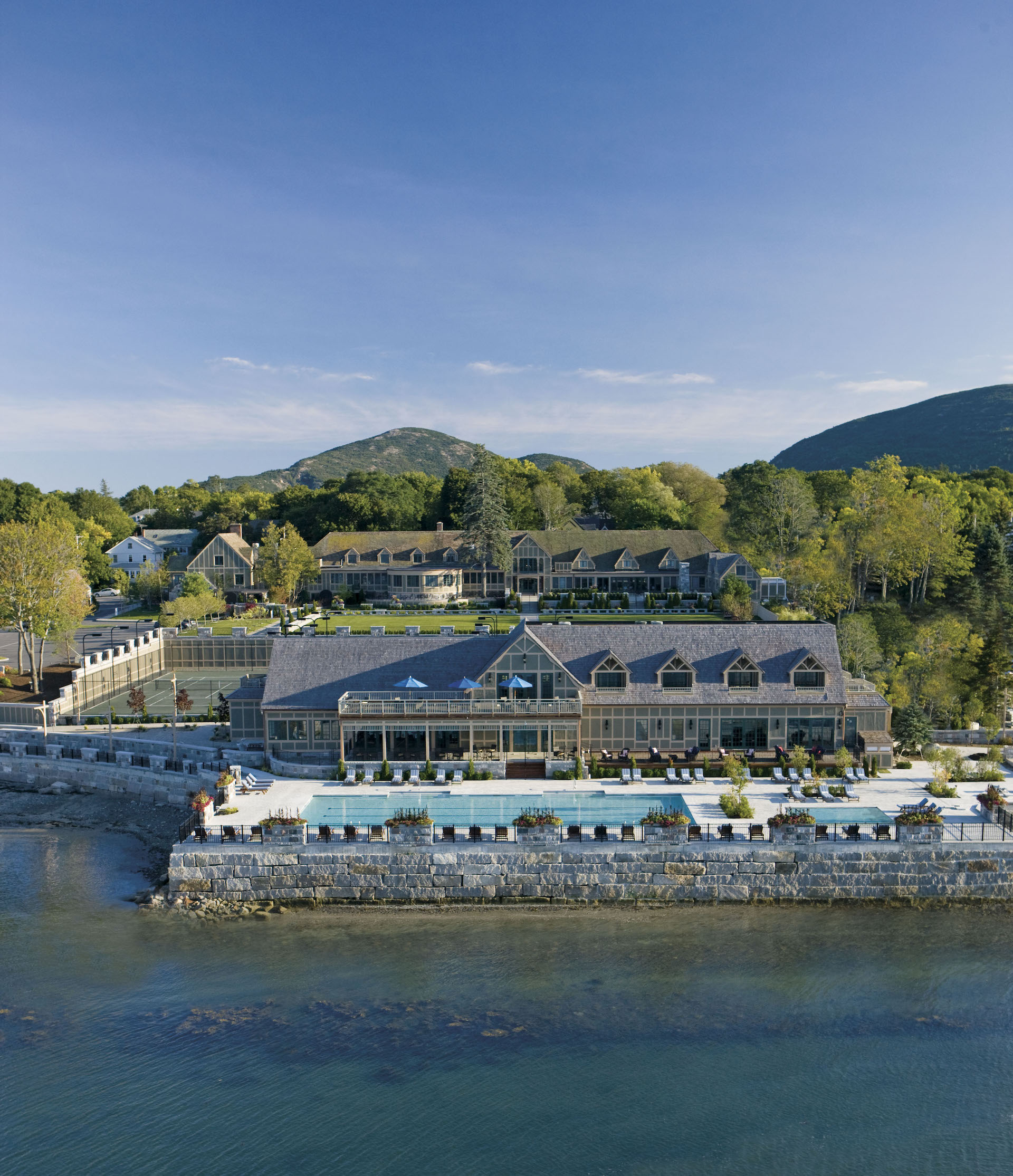 Aerial view of a large waterfront resort surrounded by lush greenery and mountains. The buildings feature gabled roofs, and theres a long rectangular pool along the edge. The sky is clear and blue.