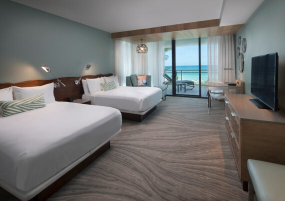 Hotel room with two queen beds, a modern design, and sea-themed decor. Theres a large window leading to a balcony with a view of the ocean, a flat-screen TV, a wooden dresser, and seating area. Soft lighting creates a cozy atmosphere.