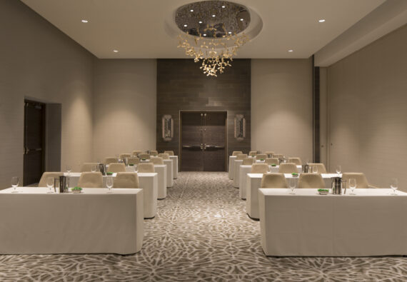 A modern conference room set up with six long tables, each paired with chairs and glassware. The room features a patterned carpet, a large chandelier on the ceiling, and neutral-colored walls with a dark wood double door at the back.