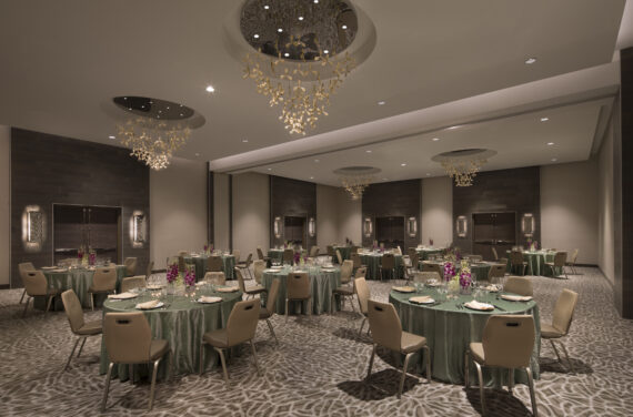 A spacious banquet hall with round tables covered in green tablecloths and set with plates and glasses. Elegant chandeliers hang from the ceiling, and purple floral centerpieces decorate the tables. The carpet has a geometric pattern.