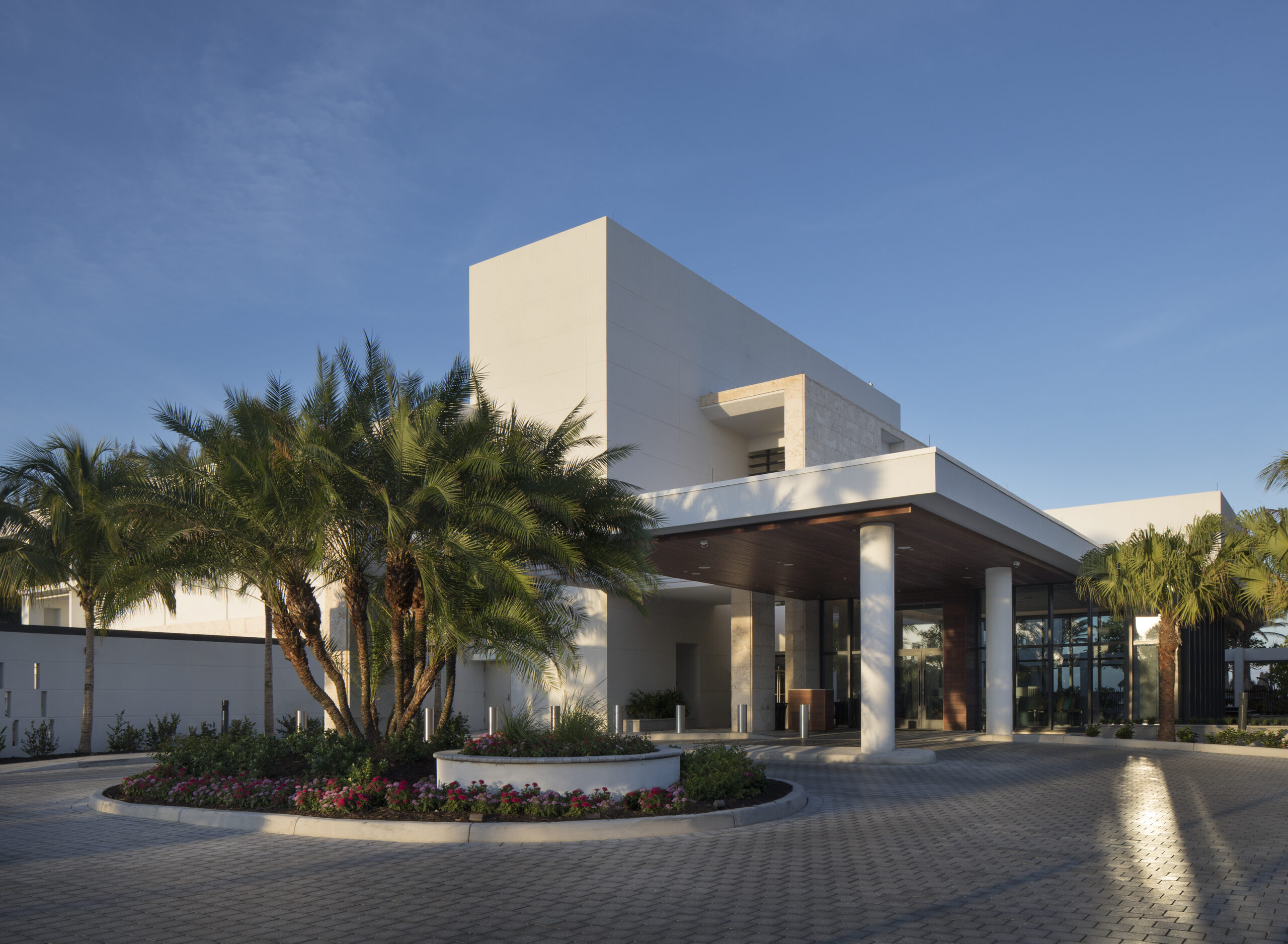 Modern white building with large windows and a shaded entrance. Surrounded by palm trees and landscaped gardens, the building sits beneath a clear blue sky. The foreground features a paved driveway.