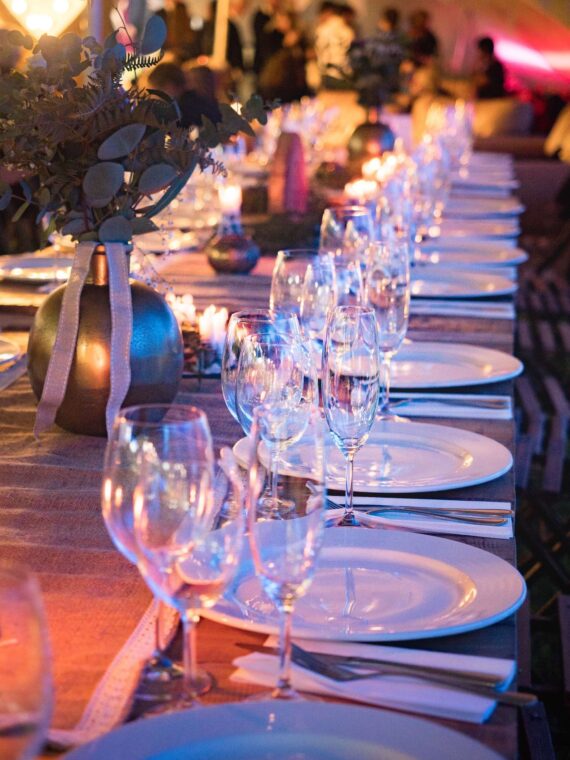 A long, elegantly set dinner table with white plates, wine glasses, and candles. Soft lighting creates a warm ambiance, and a bronze vase with greenery decorates the table. Blurred background suggests a gathering or event.