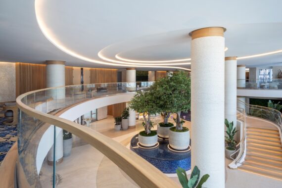 Interior of a modern building featuring a circular balcony with a glass railing. The space includes tall pillars, tree planters on a blue tiled base, and wood accents. Curved LED lighting adorns the ceiling, providing a bright and elegant atmosphere.