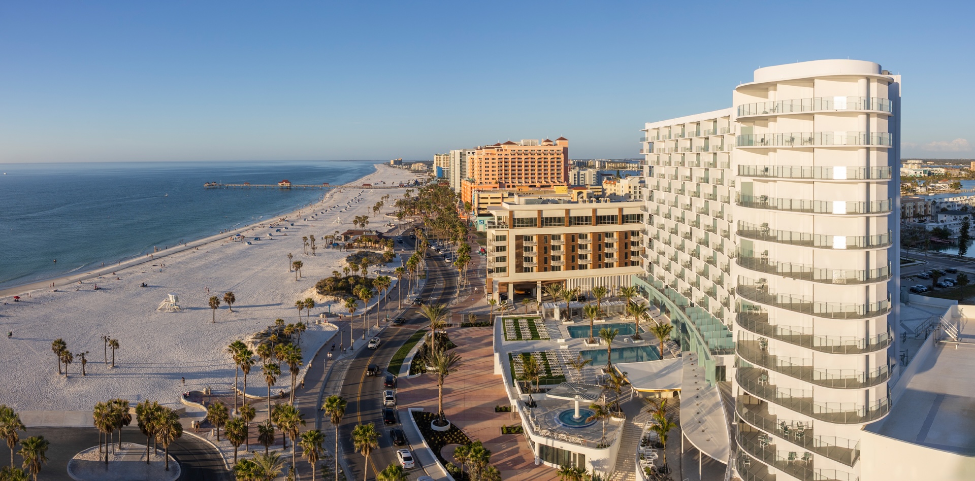 Aerial view of a coastal city featuring a wide beach with sunbathers and a long pier extending into the sea. Palm trees line the streets next to modern high-rise hotels. The ocean is calm under a clear blue sky.