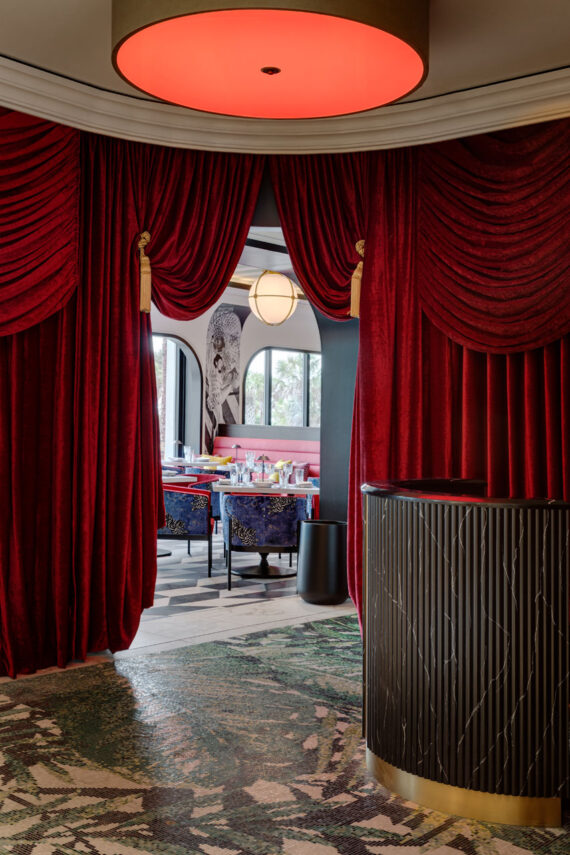 A luxurious restaurant entrance with deep red velvet curtains, a round ceiling light with a red glow, a black textured reception desk, and a stylish dining area with blue chairs and pink seating visible in the background.