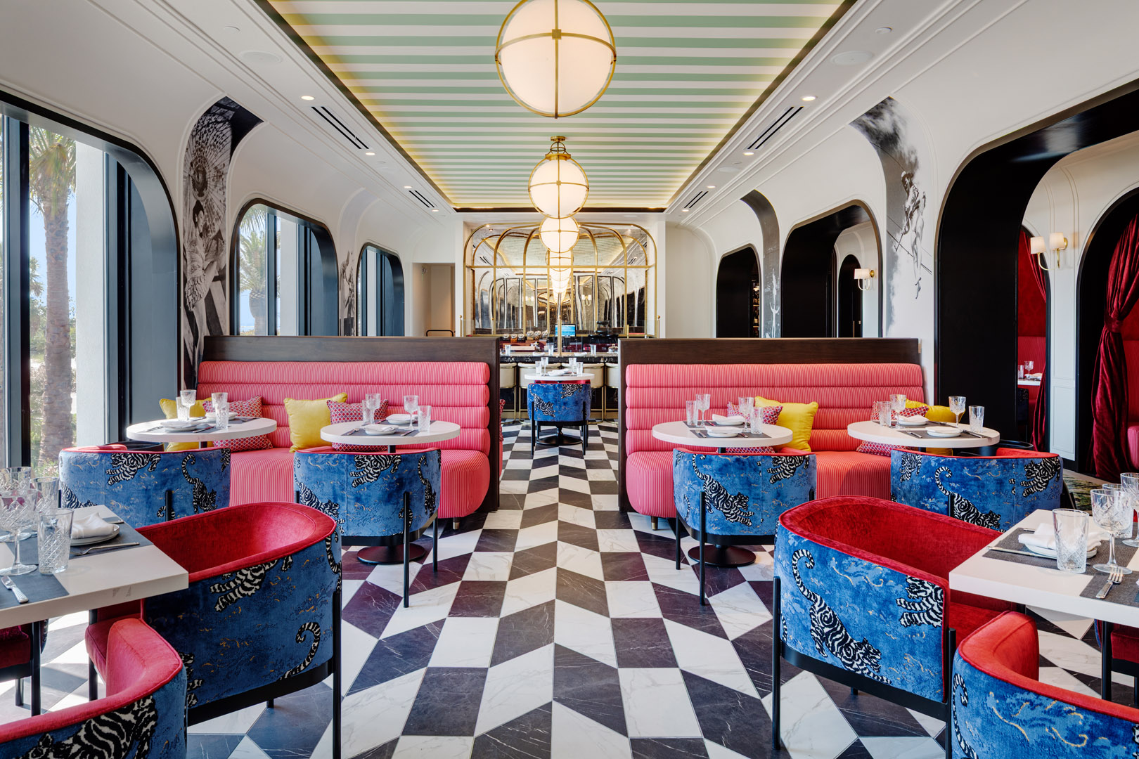 A stylish restaurant with pink booths, red and blue tiger-patterned chairs, geometric black-and-white floor tiles, large windows, and modern pendant lights hanging from a pastel green striped ceiling.