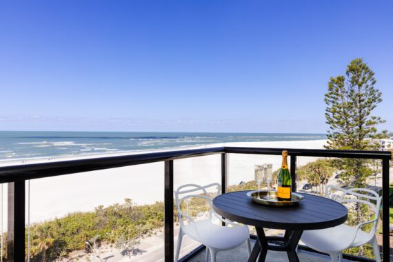 A round table with a champagne bottle and glasses sits on a balcony overlooking a sandy beach, ocean waves, and a clear blue sky, with trees visible below the balcony.
