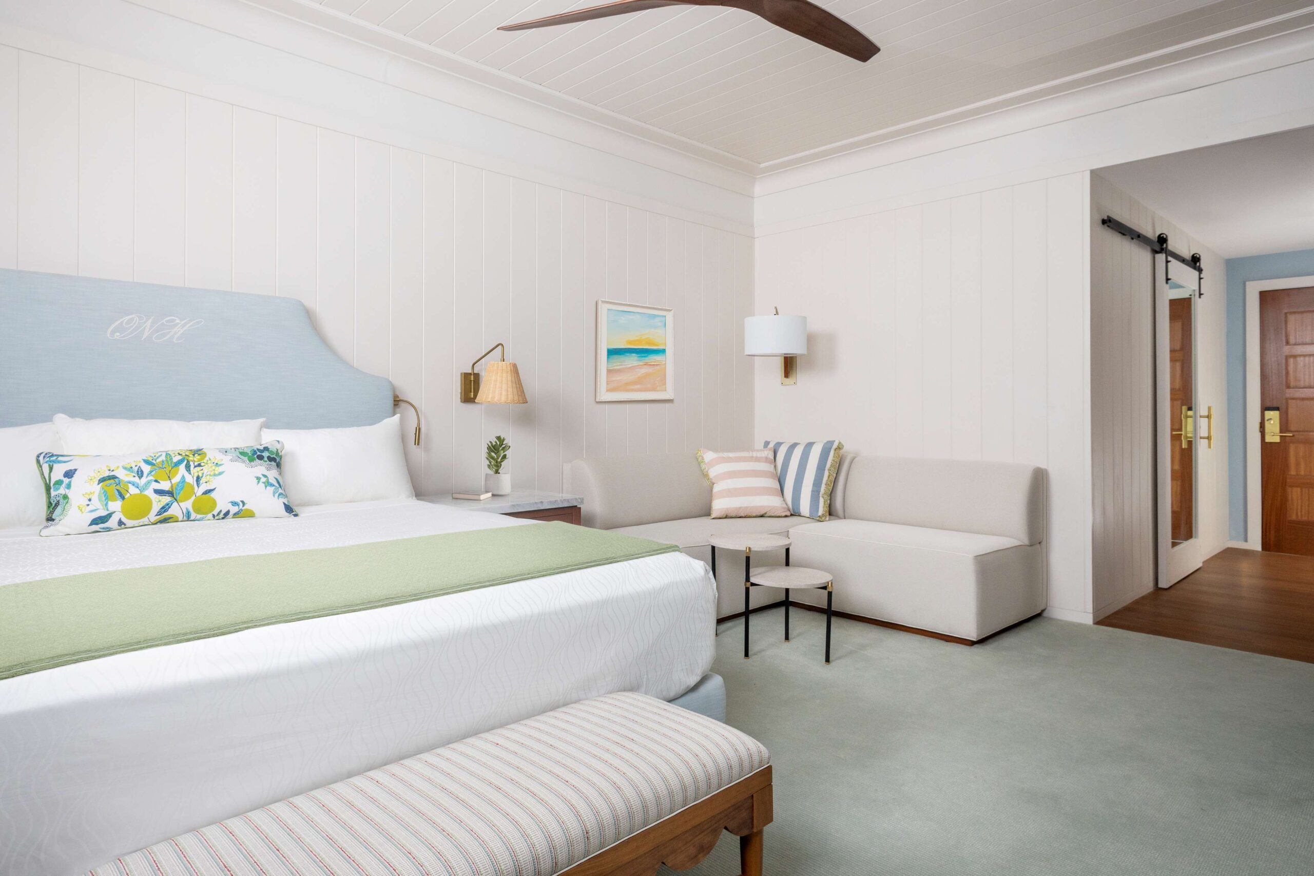 A bright, airy hotel room with a large bed featuring a blue headboard and colorful pillows. A light green runner covers the bed. Theres a beige sofa, a small coffee table, wall art, a ceiling fan, and a wooden door visible.