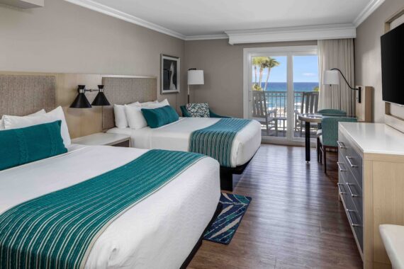A hotel room with two double beds featuring teal accents, wooden flooring, and a modern decor. A large flat-screen TV is mounted on the wall, and a sliding glass door opens to a balcony with a view of palm trees and the ocean.
