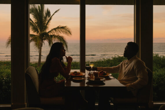 a couple eating dinner in front of a window looking out to the ocean at sunset