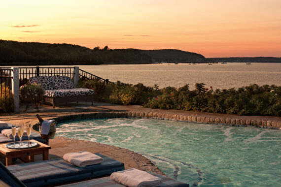 A serene view of a poolside terrace at sunset, overlooking a calm lake. The scene includes cushioned lounge chairs, rolled towels, a small table with a champagne bottle, glasses, and a scenic backdrop of forested hills and a vibrant sky.