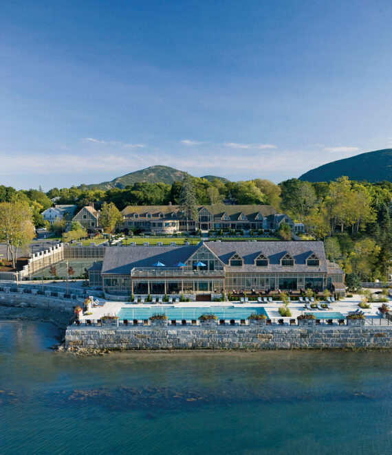 Aerial view of a coastal resort surrounded by trees and mountains. The resort features multiple buildings, a large outdoor pool, and a stone wall along the waters edge. The sky is clear and blue.
