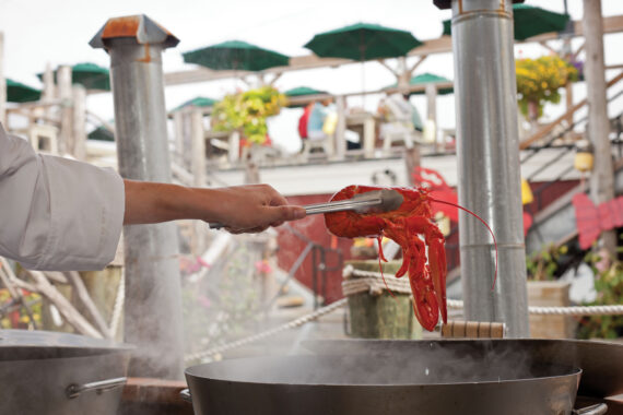 A person in a white coat uses tongs to hold a cooked lobster over a steaming pot in an outdoor setting. Green umbrellas and flowers are visible in the background, suggesting a seafood restaurant ambiance.