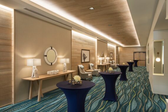 A modern hallway with elegant decor, featuring round tables draped in blue cloth, adorned with small floral arrangements and candles. Light wood accents, soft lighting, and framed art pieces create a welcoming atmosphere.