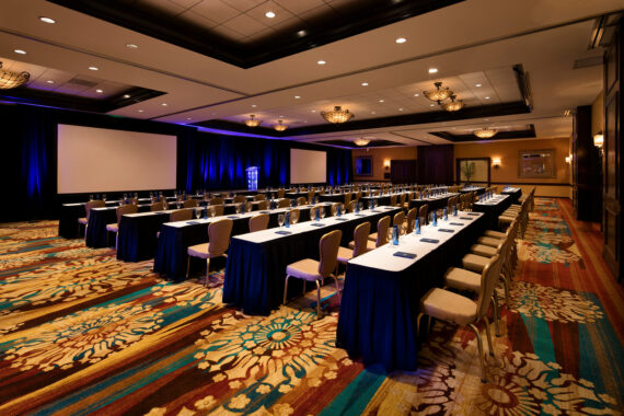 A conference room set up for an event with rows of tables and chairs facing a large projection screen. The room features colorful patterned carpet and dim overhead lighting, creating an inviting atmosphere.