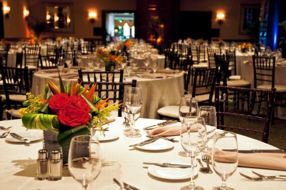 A banquet hall with elegantly set tables, featuring white tablecloths, glassware, silver cutlery, and floral centerpieces. The room is softly lit, creating a warm and inviting atmosphere.