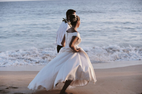 A bride and groom walk along a beach at sunset. The bride is wearing a flowing white dress, while the groom is in a white shirt and dark pants. The ocean waves gently lap at the shore beside them.