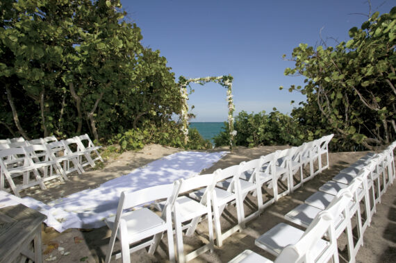 A beach wedding setup with rows of white folding chairs arranged on sandy ground. A white aisle runner leads to a floral archway overlooking the ocean, surrounded by lush greenery under a clear blue sky.