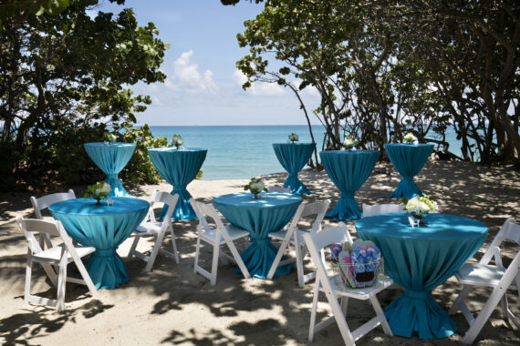 Beachside event setup with round tables and cocktail tables covered in turquoise cloths, surrounded by white folding chairs. Flower arrangements decorate the tables, and the ocean is visible in the background under a clear, sunny sky.