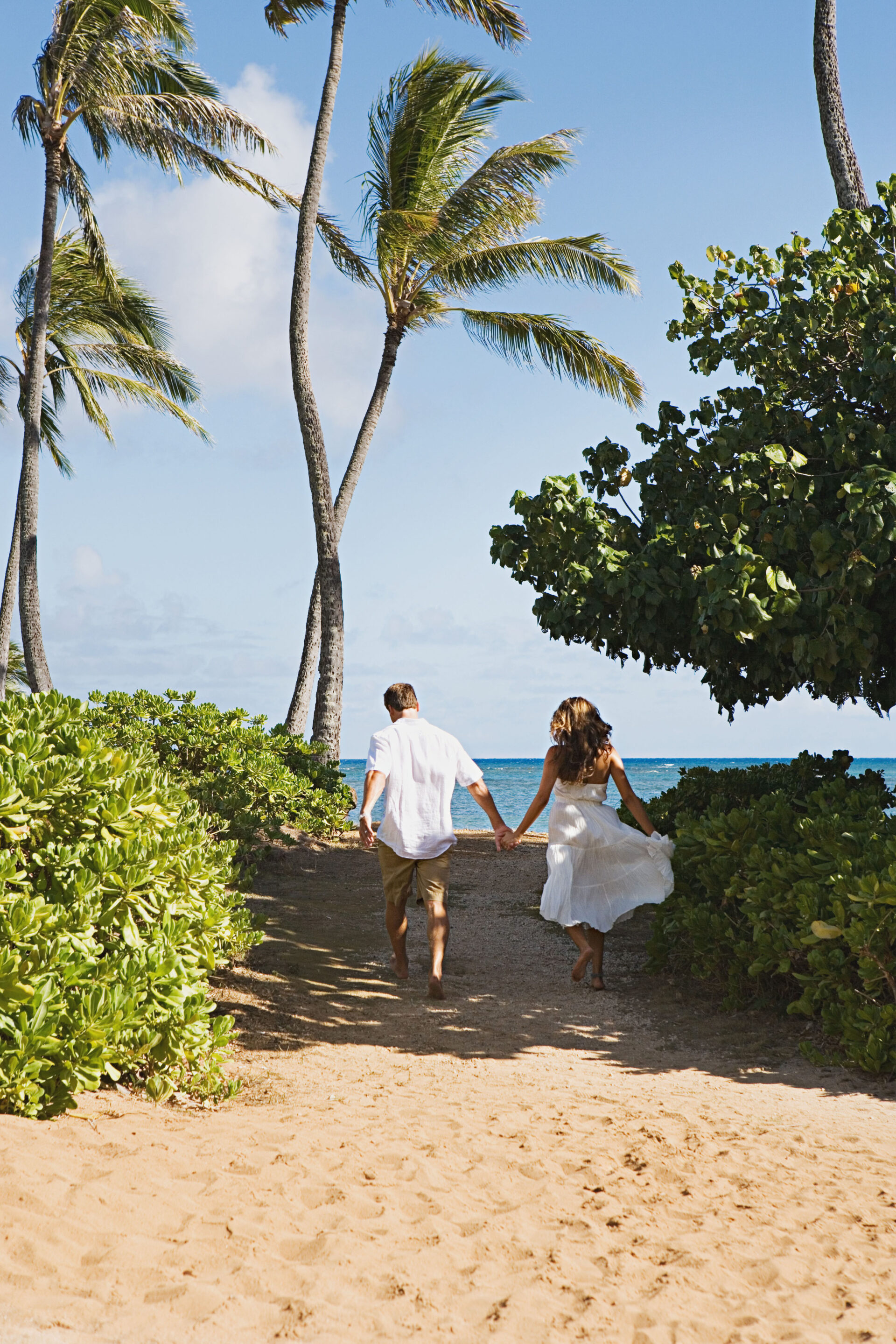 A couple holding hands runs towards a beach, surrounded by lush greenery and tall palm trees. The sky is clear and blue, evoking a sense of joy and freedom.