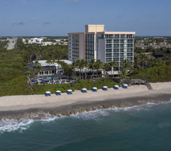 A beachfront hotel with a light pink facade rises above palm trees and a sandy beach lined with blue and white umbrellas. The ocean waves gently lap the shore under a clear blue sky.