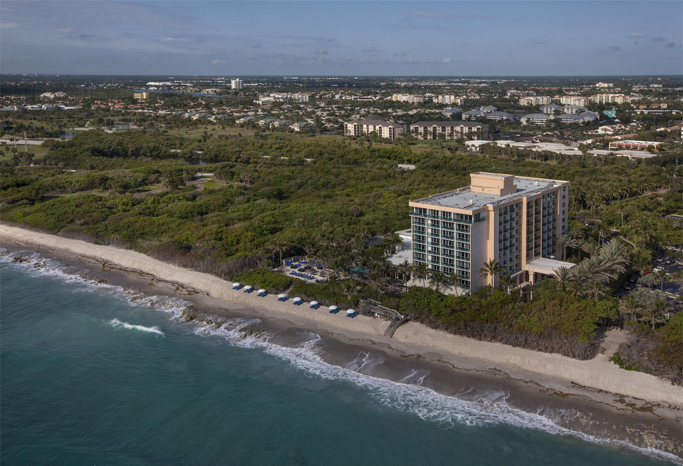 Aerial view of a beachfront hotel surrounded by lush greenery. The hotel is adjacent to a sandy beach with waves gently reaching the shore. Sun loungers with blue umbrellas are arranged on the sand. Urban development is visible in the background.