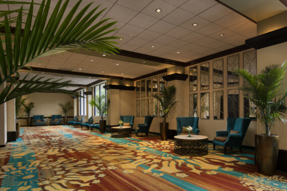 A spacious lobby with vibrant, floral-patterned carpet, blue armchairs, and round tables adorned with potted plants. Large windows and decorative mirrors on the walls enhance the spacious, well-lit atmosphere.